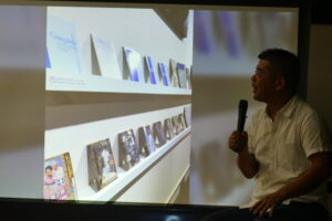 吉澤健さんの作品が展示されている写真を見ながら話す館長